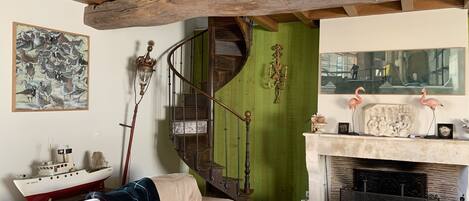 A cozy living room with a wooden ceiling, a green wall, a fireplace with decorative items, a staircase, and a large abstract painting.