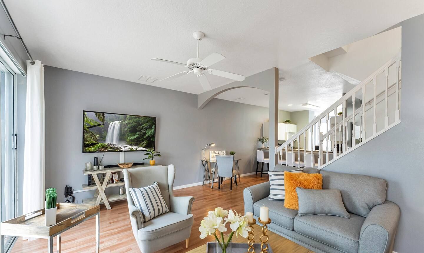 Un salon moderne avec un canapé gris, un fauteuil  à rayures et une table basse en bois sur laquelle est posé un vase de fleurs. Un téléviseur à écran plat est fixé au mur, et un ventilateur de plafond est visible.