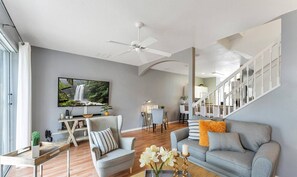A modern living room with a grey sofa, a striped armchair, and a wooden coffee table with a vase of flowers. A flat-screen TV is mounted on the wall, and a ceiling fan is visible.