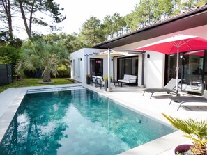 Un área de piscina exterior moderna con una piscina cristalina, sillas de descanso y una zona de estar cubierta con una sombrilla roja.
