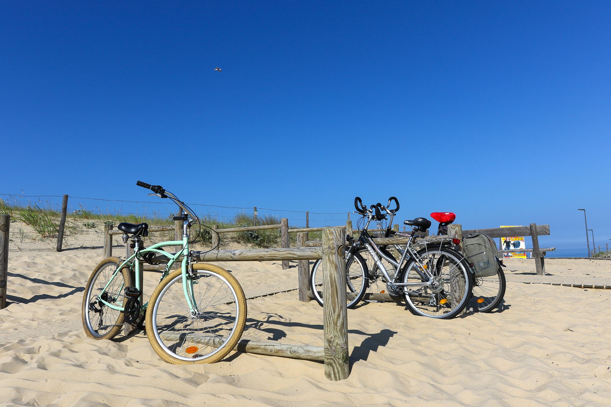 Bicicletas estacionadas en una playa arenosa con una cerca de madera y un cielo azul despejado al fondo.
