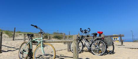 Bicicletas estacionadas en una playa arenosa con una cerca de madera y un cielo azul despejado al fondo.