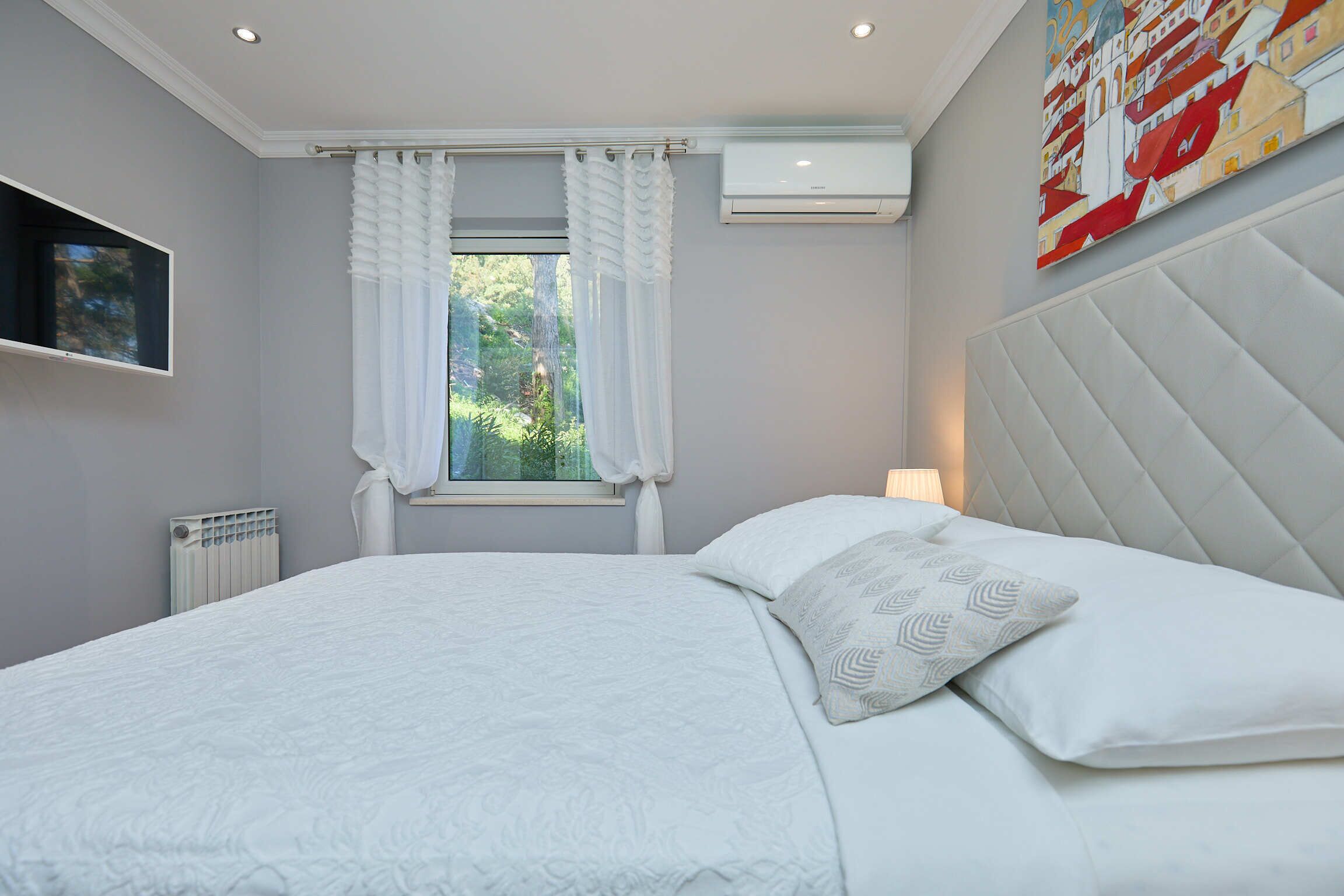Una camera da letto con un letto, una finestra con tende, un condizionatore, un termosifone, una TV a muro e un quadro appeso.