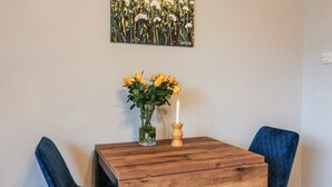 A wooden table with a vase of yellow flowers and a candle, flanked by two blue chairs, in a room with a floral painting on the wall. - Cornerways, 8 North Lane (York)