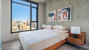 Een moderne slaapkamer met een groot raam, een bed met witte lakens, twee abstracte schilderijen en een nachtkastje met een lamp.