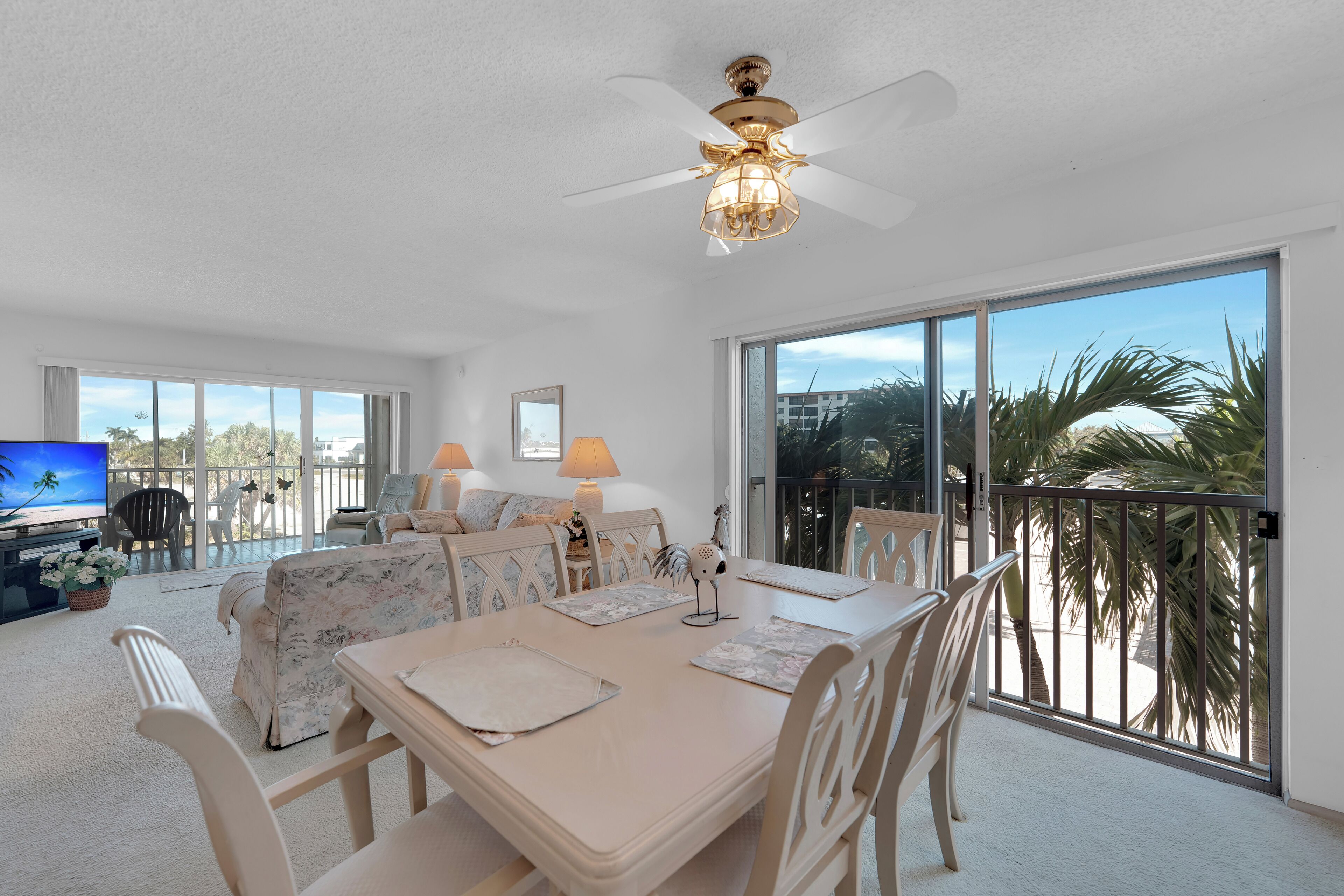 A spacious living area with a dining table, chairs, a ceiling fan, and large windows offering a view of palm trees and a building.