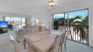 A spacious living area with a dining table, chairs, a ceiling fan, and large windows offering a view of palm trees and a building. - HELLO SUNSHINE! FUN IN THE SAND! FAMILY ISLAND VACATION ON A BUDGET! (Fort Myers Beach)
