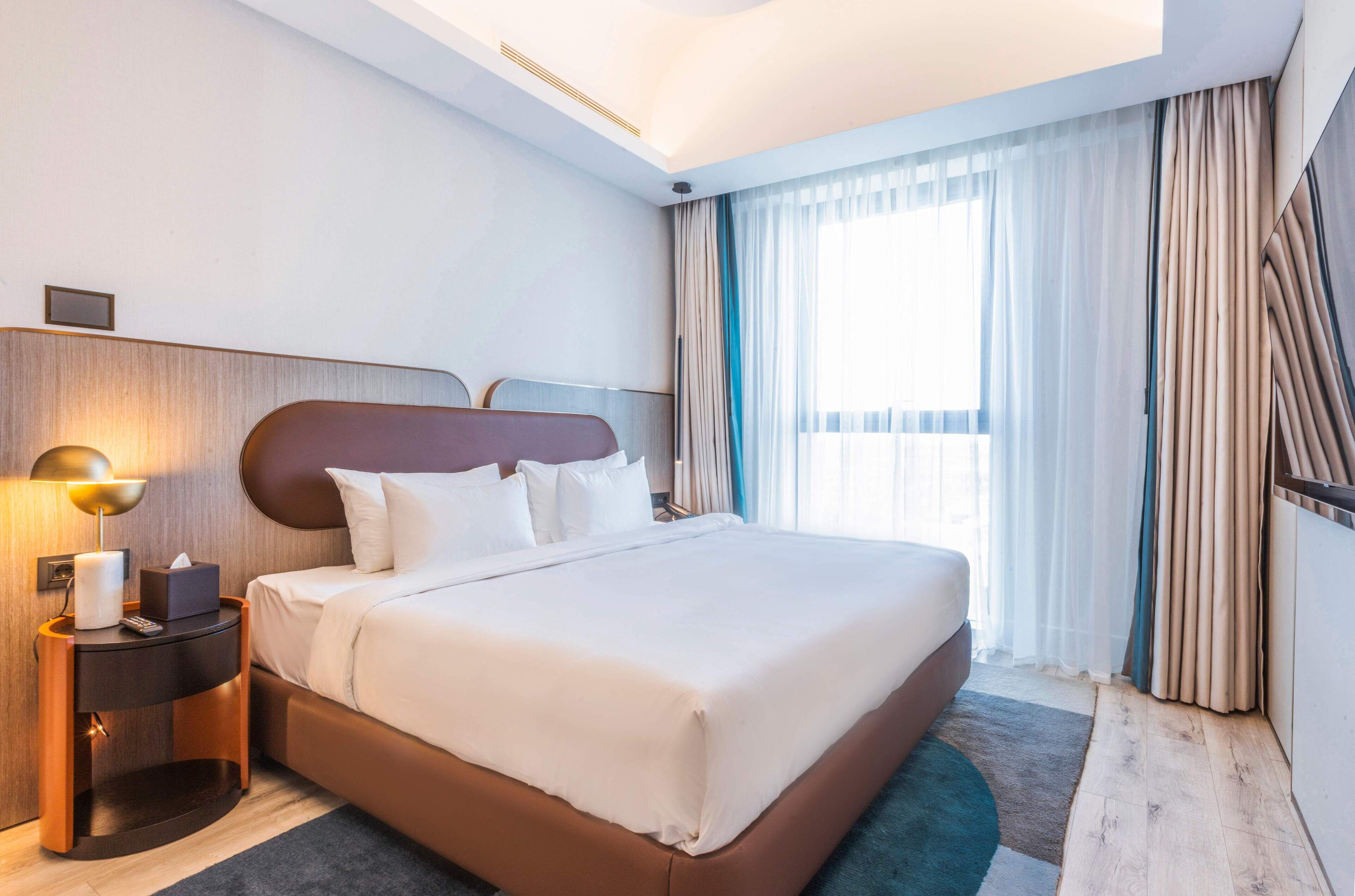 Una habitación de hotel moderna con una cama grande, una mesita de noche con una lámpara y vistas al exterior a través de cortinas translúcidas.