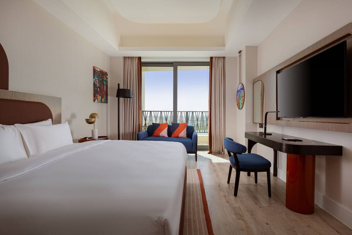 A hotel room with a large bed, a desk with a chair, a TV, and a balcony with curtains.