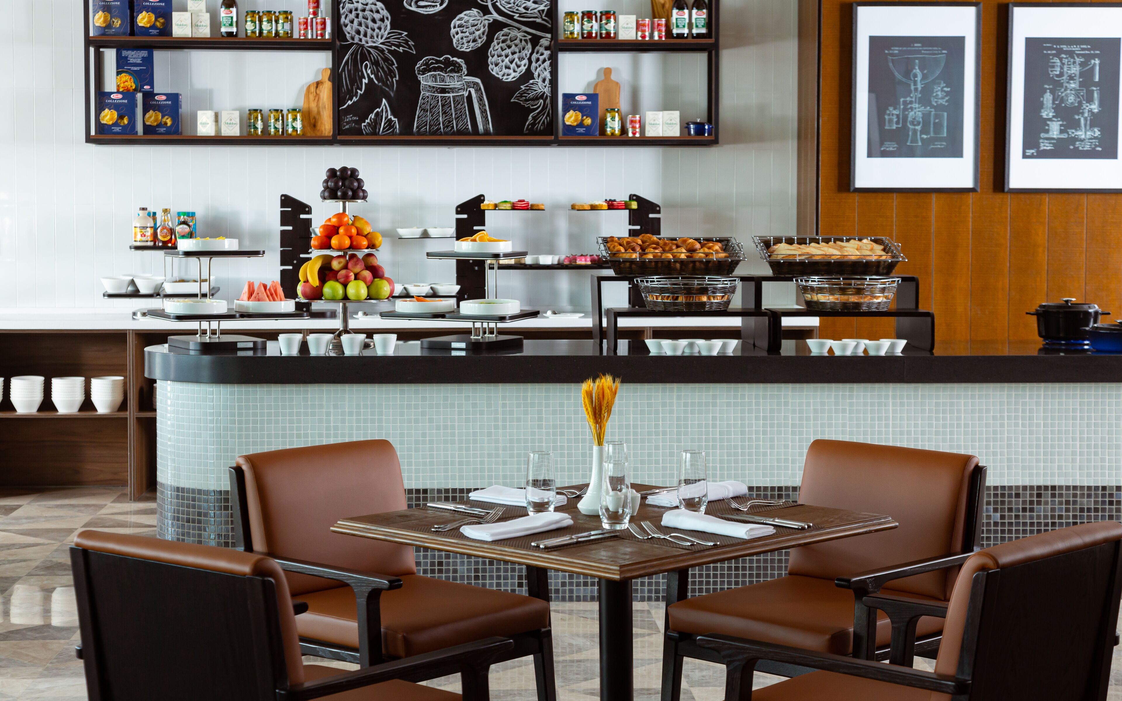 A restaurant with a buffet setup, including fruits, pastries, and condiments. There are two tables with chairs, and the decor includes a chalkboard with fruit illustrations.
