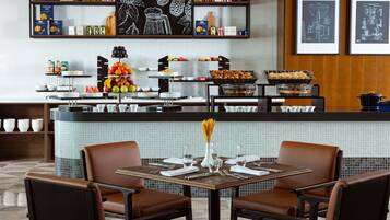 A restaurant with a buffet setup, including fruits, pastries, and condiments. There are two tables with chairs, and the decor includes a chalkboard with fruit illustrations.
