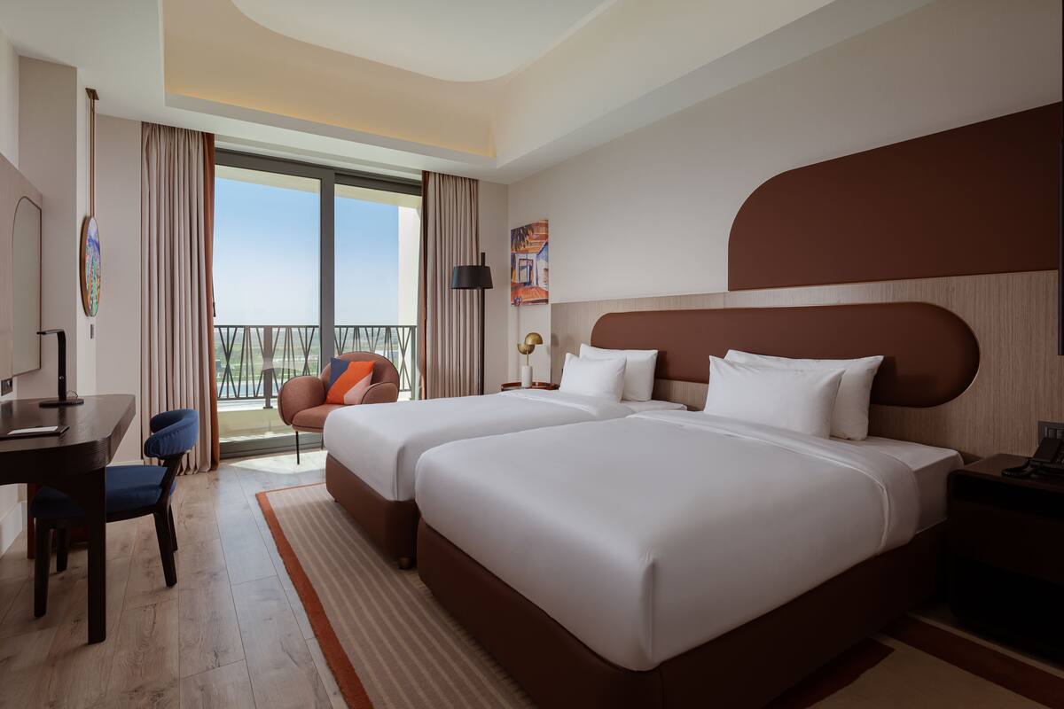 A hotel room with two beds, a desk, a chair, a balcony with a view, and a painting on the wall.