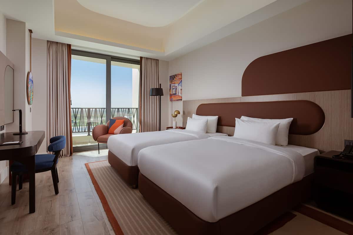 A hotel room with two beds, a desk, a chair, a balcony with a view, and a painting on the wall.