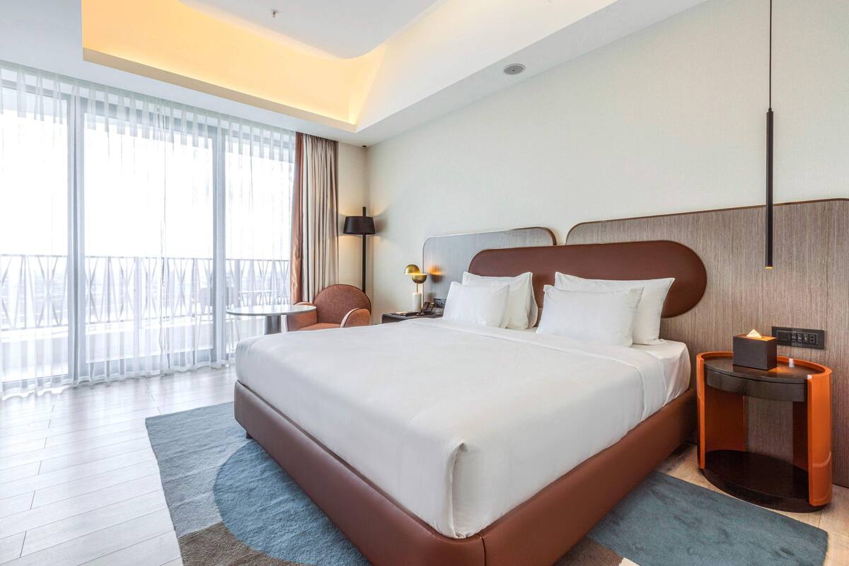 A modern hotel room with a large bed, a bedside table with a lamp, and a view of the outdoors through large windows.