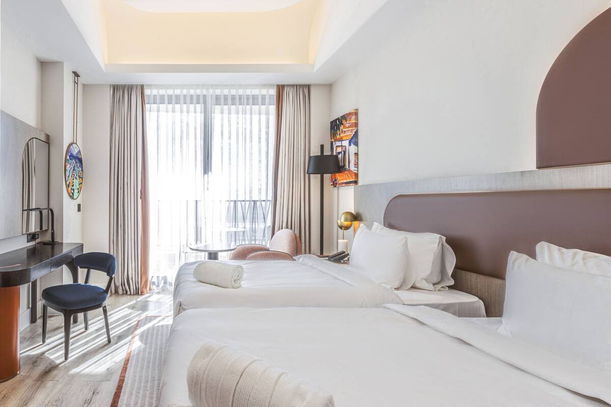 A hotel room with two beds, a desk, a chair, a TV, and a window with curtains.