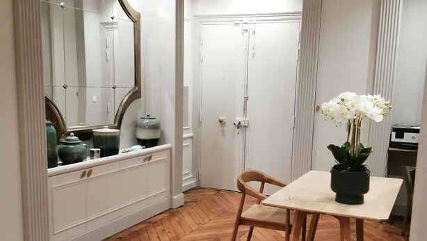 A room with a herringbone wooden floor, a white vanity with a mirror, a small table with a potted orchid, and a closed door. - Etoile-Champs Elysée (Paris)