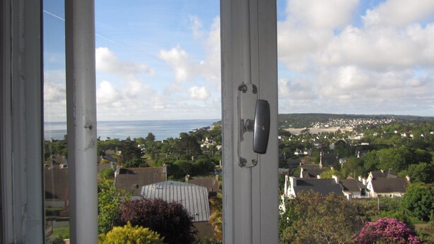 A view through a window with a metal handle, overlooking a residential area with houses and a body of water in the distance. - Villa in the center of Crozon (Crozon)