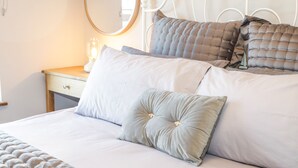 Una cama bien hecha con edredón blanco, almohadas grises y blancas, y un espejo redondo sobre una mesita de noche con una lámpara.