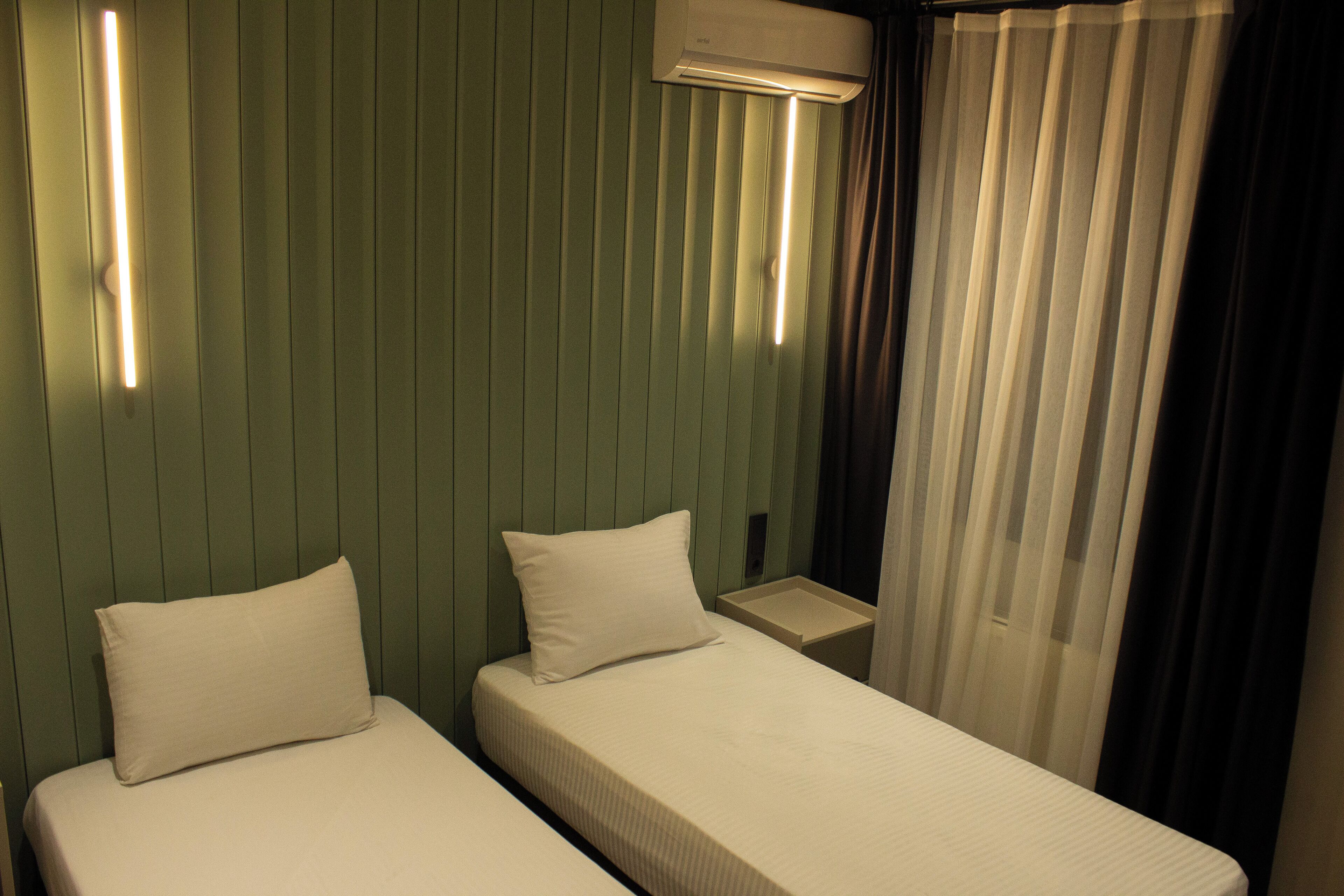 Quarto de hotel com duas camas de solteiro, mesa de cabeceira, ar condicionado fixado na parede e janela com cortinas.