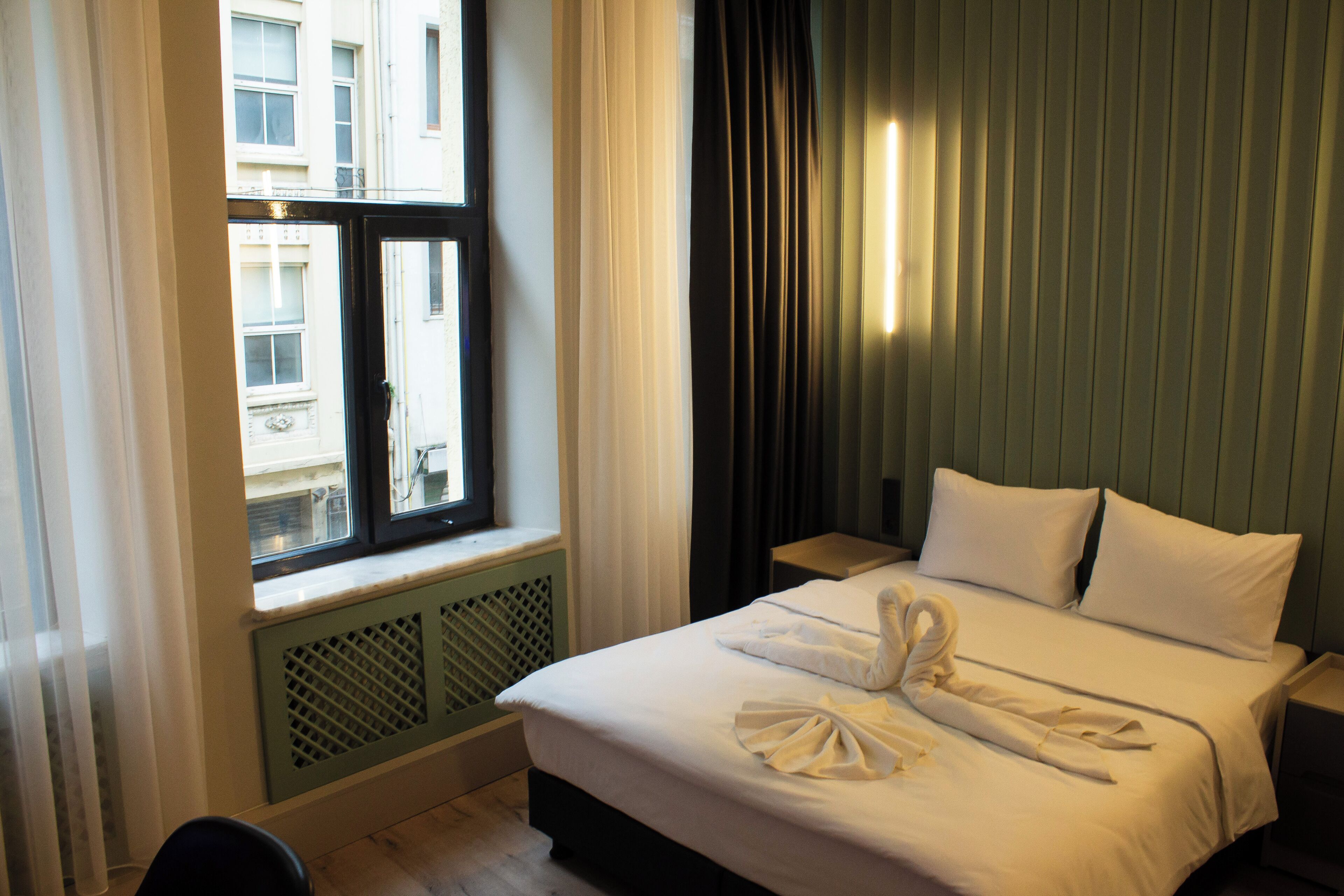 A hotel room with a bed, a window with curtains, a radiator, and a nightstand with a lamp.