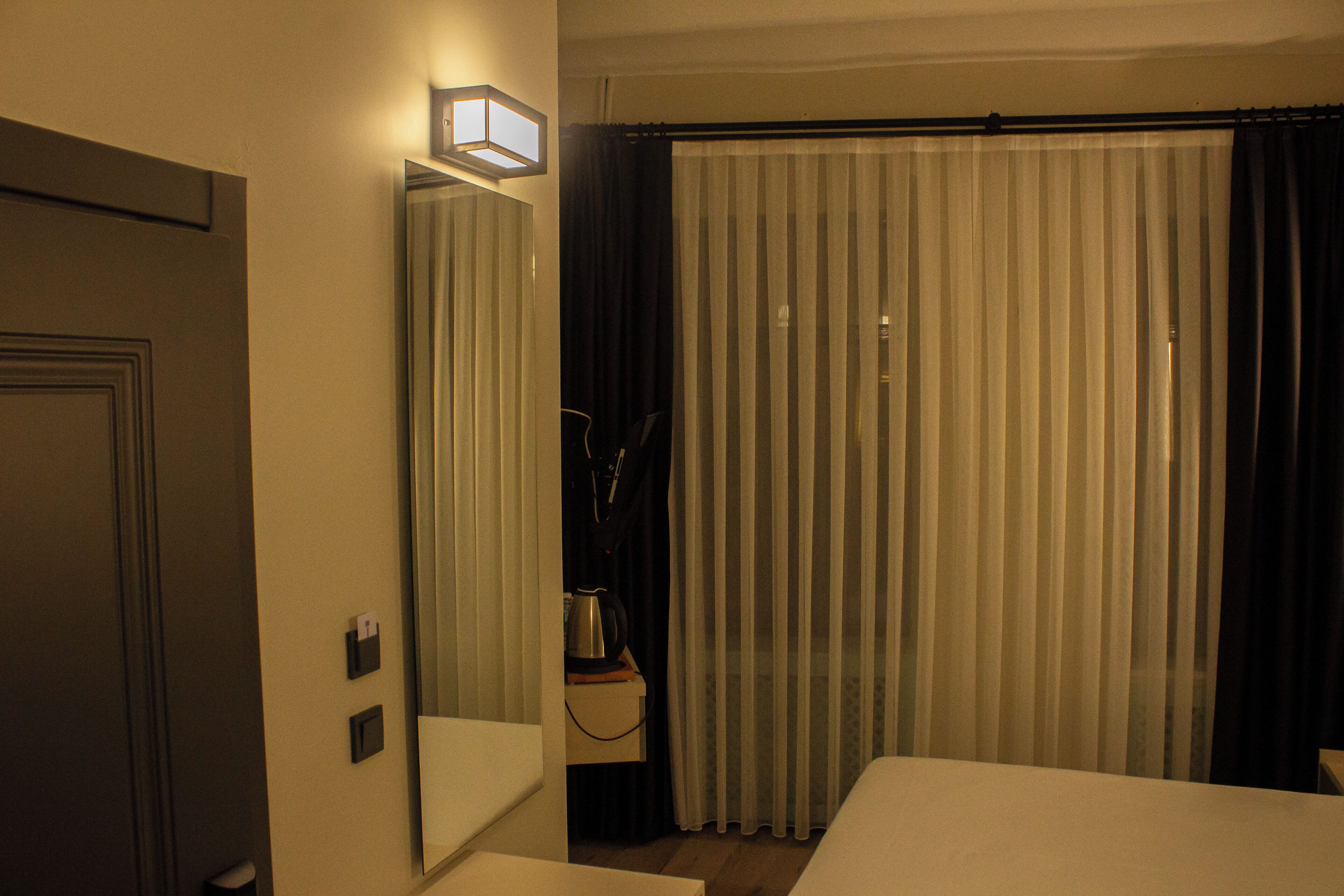 A hotel room with a bed, a bedside table, a mirror, a curtain, and a wall-mounted light fixture.