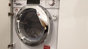 A front-loading washing machine with a control panel, a safety lock, and a red sticker with contact information. - Stylish detached studio with a wet room, Satellite TV & other amenities 😀 (Bristol)
