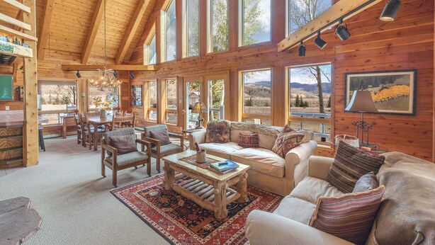 A spacious log cabin interior with a dining area, a living room with a sofa and armchairs, and large windows offering views of the outdoors. - Custom Log Home-Pet-Friendly-Rio Grande River Access-Breathtaking Views (Creede)