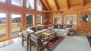 A spacious living room with a high ceiling, wooden walls, and large windows offering a view of the outdoors. - Custom Log Home-Pet-Friendly-Rio Grande River Access-Breathtaking Views (Creede)