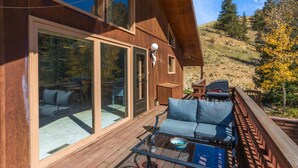 A wooden deck with a glass door leading to a patio area, a seating area with a table, and a view of a hilly landscape. - Spacious Retro Mountain Getway - ATV Parking - Amazing Views (Creede)