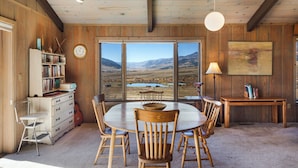 A dining area with a large table and chairs, a bookshelf, a clock, and a view of a landscape through large windows. - Spacious Retro Mountain Getway - ATV Parking - Amazing Views (Creede)