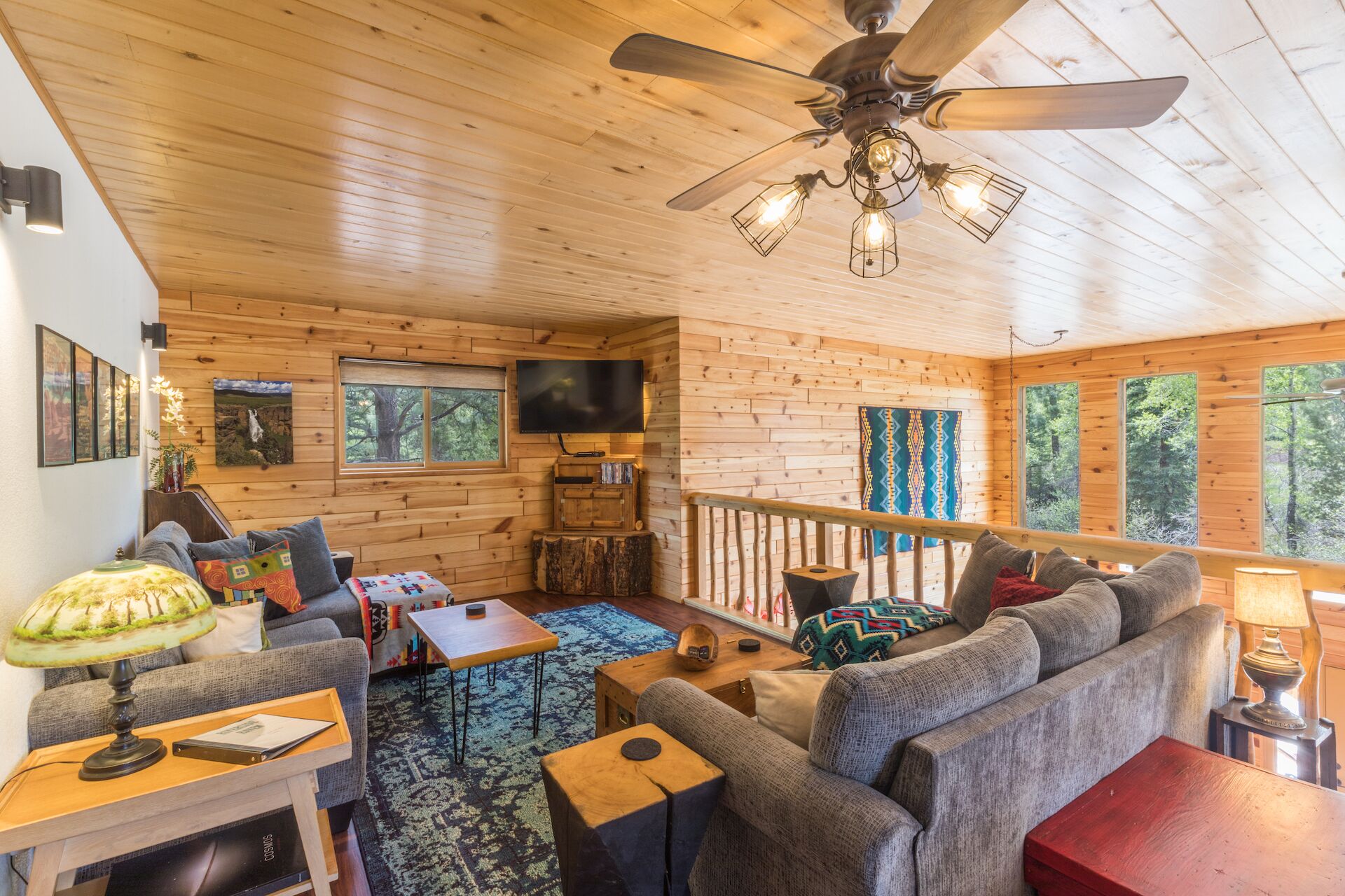 L’intérieur chaleureux d’un chalet avec un plafond en bois, un ventilateur de plafond, une télévision, un canapé, une table basse et une fenêtre donnant sur la forêt.