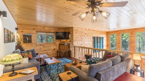 Ein gemütliches Blockhäuschen-Interieur mit Holzbalkendecke, Deckenventilator, Fernseher, einer Couch, einem Couchtisch und einem Fenster mit Blick auf den Wald.