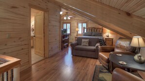 Et hyggeligt interiør i en hytte med trævægge, et sovehems med seng og skrivebord, en opholdsstue med sofa og læderstol samt et spisebord med lampe.