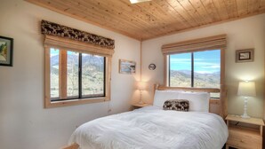 A bedroom with a wooden ceiling, two windows with mountain views, a bed with white bedding, and a nightstand with a lamp. - Secluded Mountain Cabin - Pet Friendly - National Forest Access (Creede)