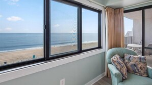 Quarto com uma grande janela com vista para a praia, uma poltrona azul-turquesa com almofadas estampadas e vista para o oceano.
