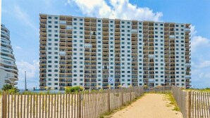 Um hotel moderno com várias varandas, um caminho de areia que leva à entrada e um céu claro.
