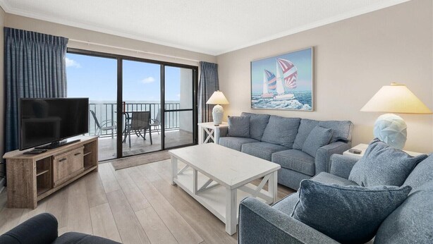 Sala de estar moderna com televisão de tela plana, mesa de centro branca, sofá cinza e varanda com vista para o mar.