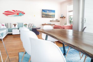 A modern living room with a wooden dining table, white chairs, a teal sofa, and a red sofa. There are wall art pieces and a window with blinds.