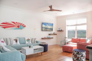 A spacious living room with a light blue sofa, a white coffee table, and a pink ottoman. There are wooden shelves on the wall and a ceiling fan.