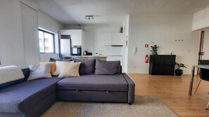 A modern living room with a grey sectional sofa, a small kitchen area with white cabinets, and a wooden floor.