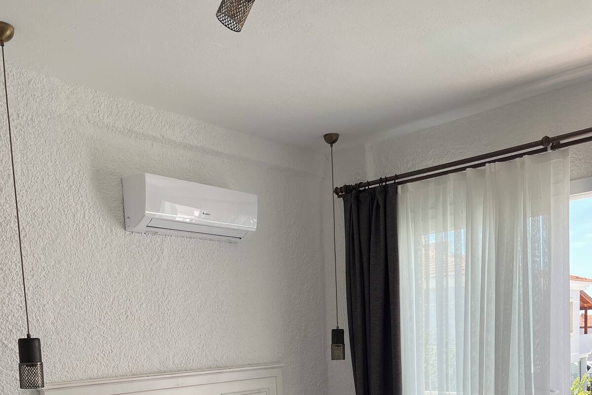 A bedroom with a bed, white walls, a ceiling fan, an air conditioning unit, a nightstand, a bedside lamp, and a window with curtains.
