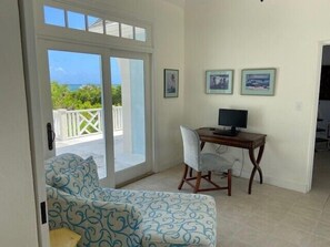 Una habitación con puerta de vidrio corrediza que da a un balcón, un escritorio con computadora, un sofá estampado y cuadros en la pared.