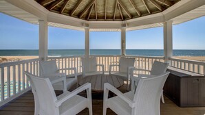 Een overdekt terras met witte rieten stoelen en een centrale tafel, met uitzicht op een strand.