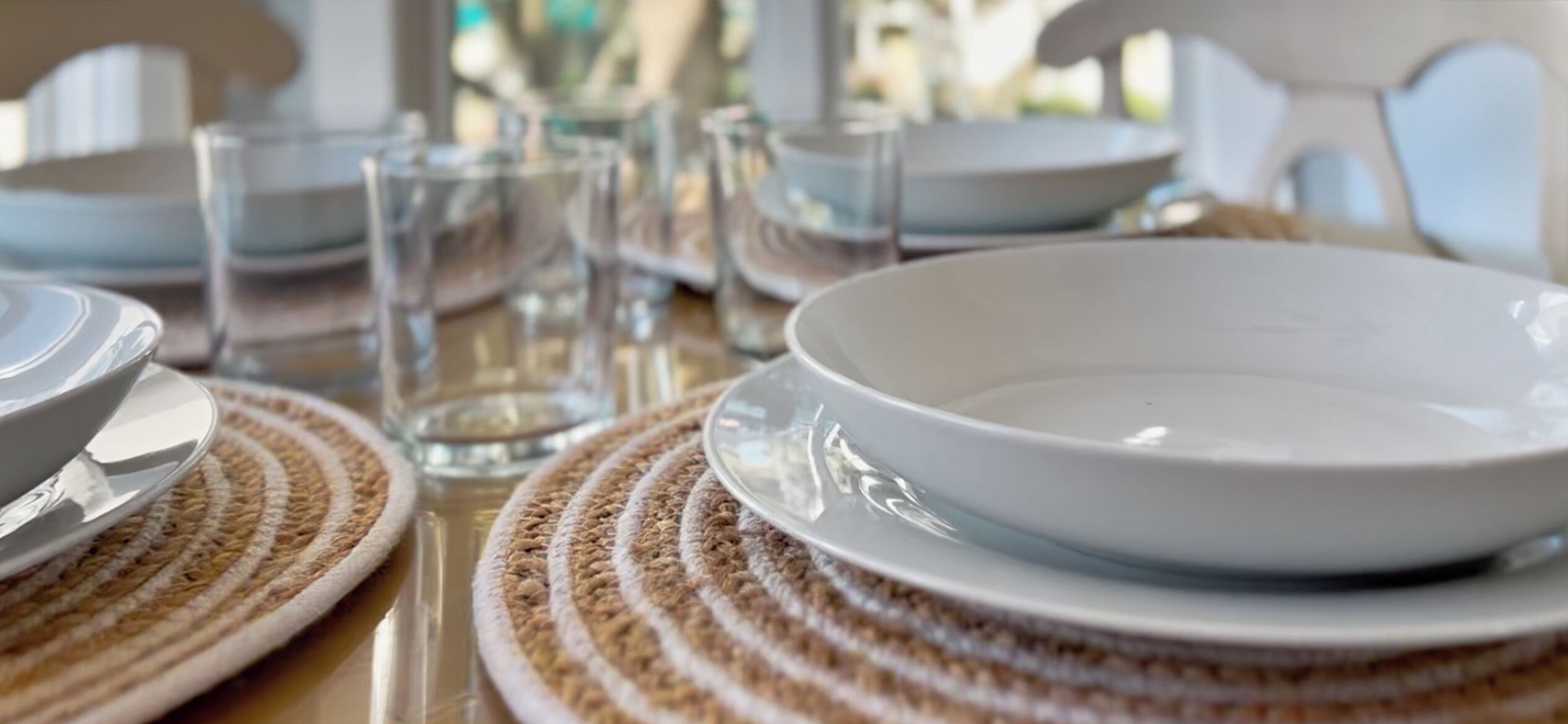 Une table à manger avec des assiettes blanches, des verres transparents et des sets de table en fibres tressées.