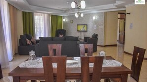 A modern living room with a sofa, dining table, and chairs. There is a television on the wall and a ceiling fan. - Stay.Plus Mount Kenya Road Nyali (Mombasa)