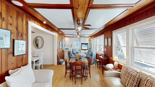 A spacious living area with wooden paneling, a ceiling fan, a dining table with chairs, and a sofa.