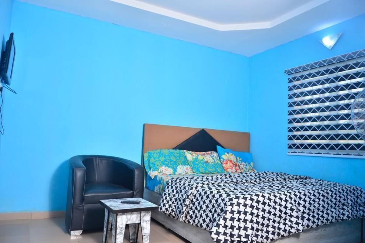 Un dormitorio con cama, una silla, una mesita y un televisor montado en la pared.