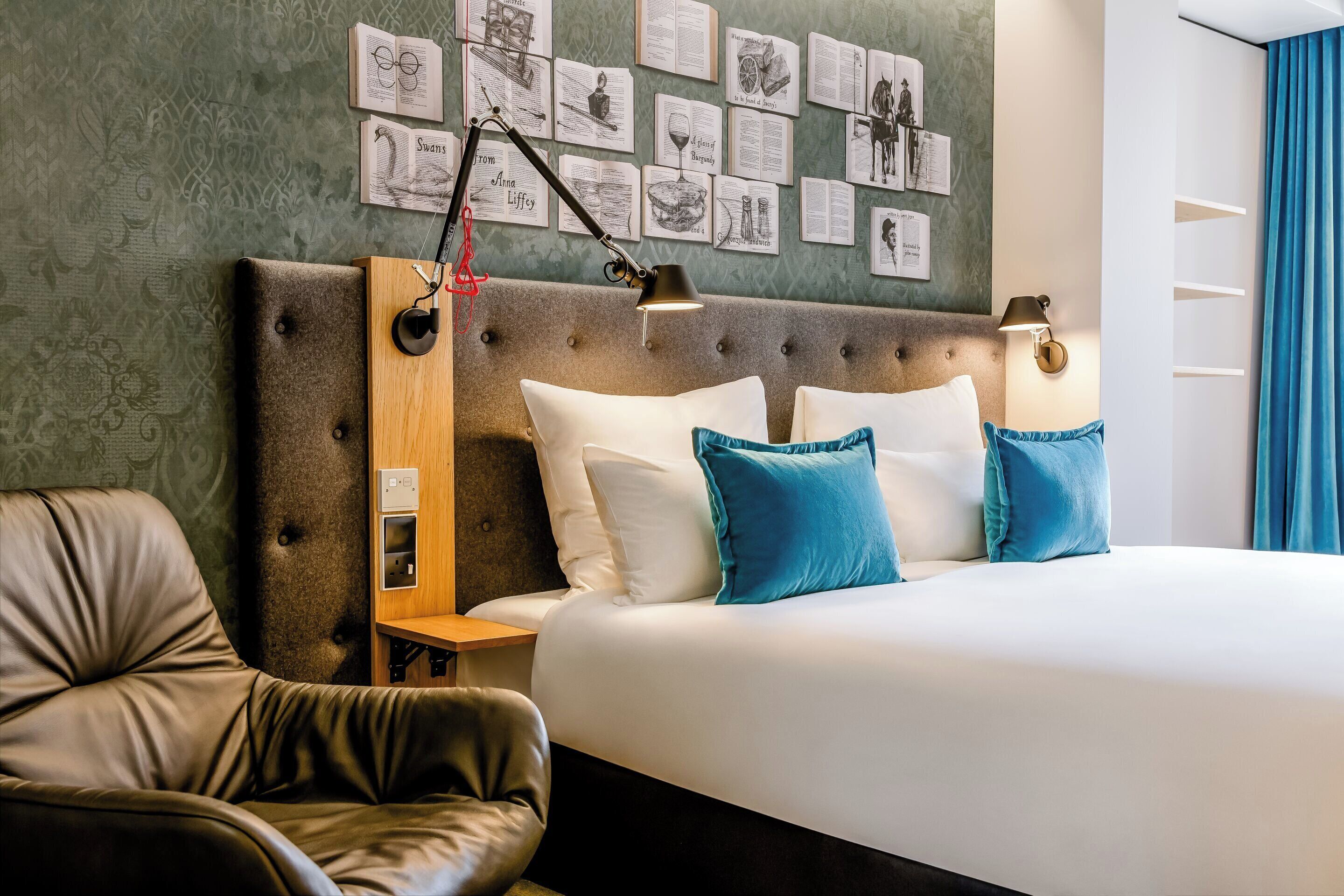 A hotel room with a tufted headboard, a bed with white and blue pillows, a leather armchair, a nightstand with a lamp, and a wall decorated with framed pictures.