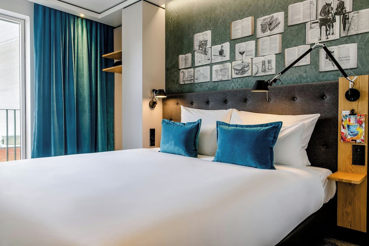 A neatly made bed with white linens and blue pillows, a headboard with a tufted design, a wall with framed pictures, and a window with blue curtains.