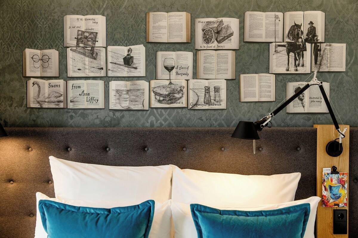 Une chambre à coucher avec un lit, des oreillers, une table de chevet avec une lampe, et un mur décoré de livres ouverts et d’illustrations.